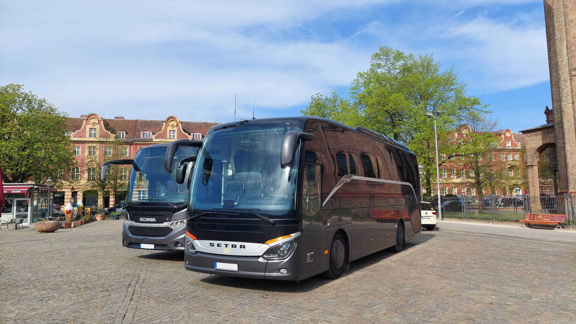 Zwei moderne Reisebusse für Fahrservice für Gruppen stehen auf einem gepflasterten Platz nahe historischen Gebäuden und einer Kirche mit hohem Uhrturm unter leicht bewölktem Himmel in Potsdam. Ideal für Busfahrten, Gruppenreisen und Vereinsfahrten in Berlin & Brandenburg.