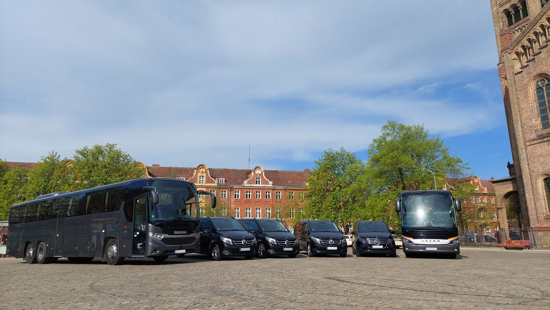 Eine Reihe schwarzer Mercedes-Benz-Vans und Busse stehe auf einem gepflasterten Platz vor einem traditionellen roten Backsteingebäude unter blauem, leicht bewölktem Himmel.