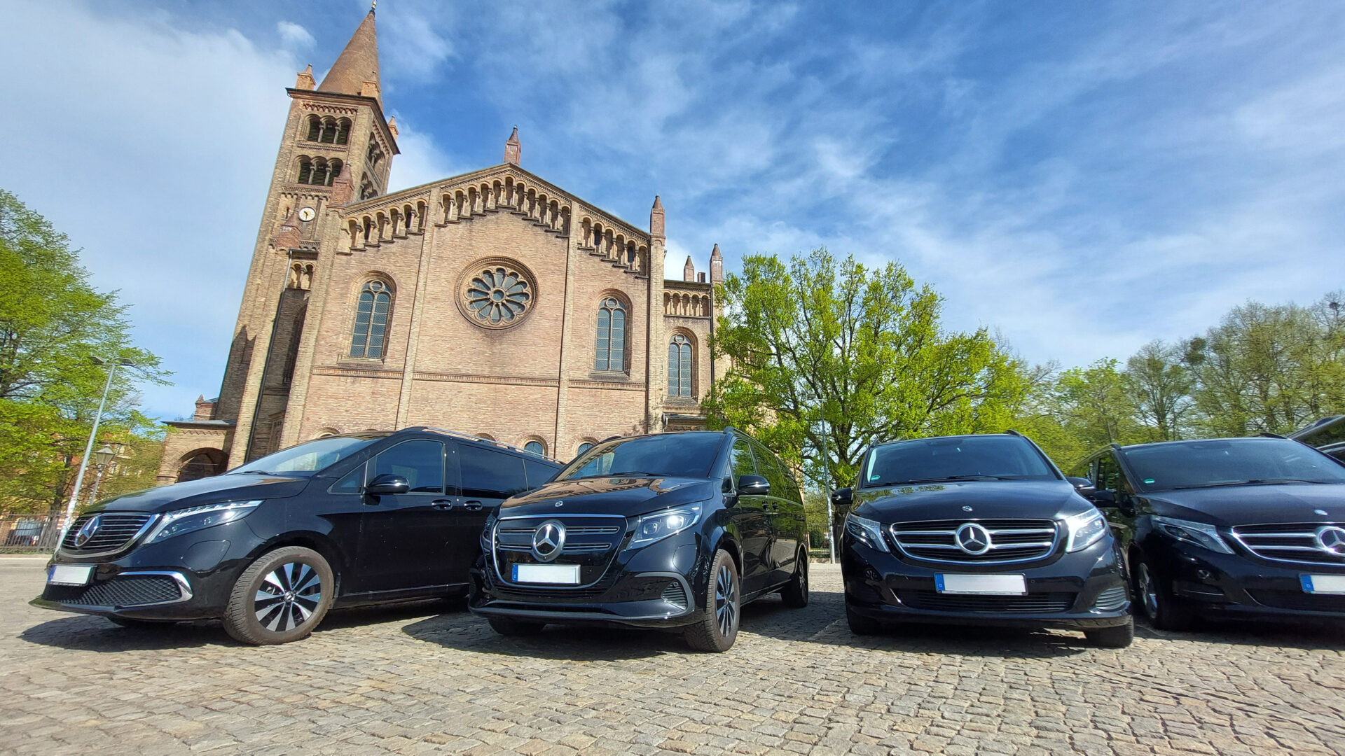 Mehrere schwarze Mercedes-Benz-Vans stehen auf einem gepflasterten Platz vor einer großen historischen Backsteinkirche unter blauem, leicht bewölktem Himmel.